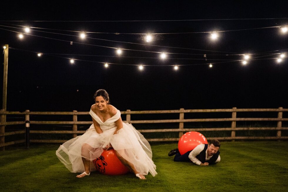 Funny wedding photo: Bride bouncing on hopper ball, groom lies on the grass with a hopper ball. Outdoor wedding reception.